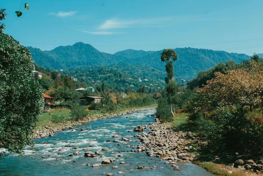 A shallow river running over stones through a wooded valley with houses on the far bank